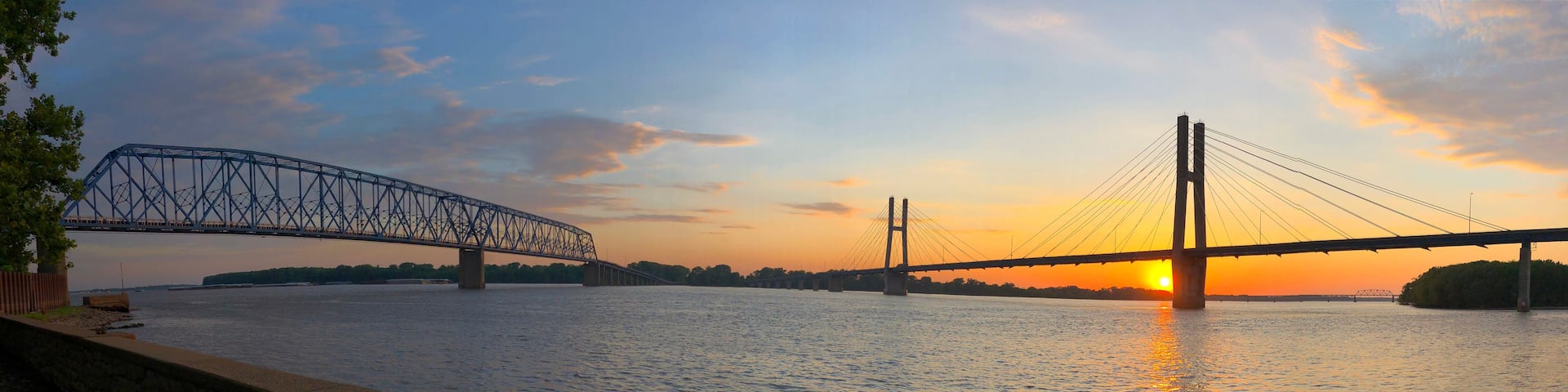 A sunset over a river with two bridges, one with a truss design and the other with cables.