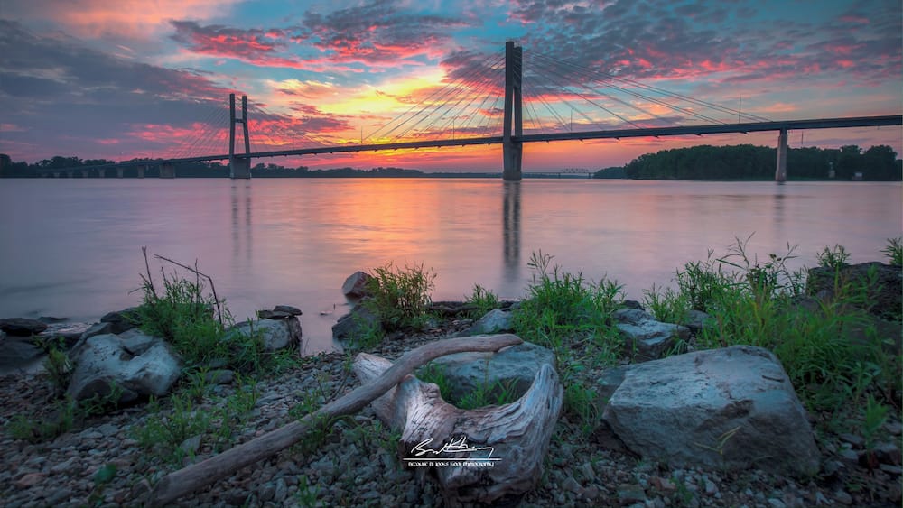 A suspension bridge at sunset with a calm river in the foreground and a colorful sky.