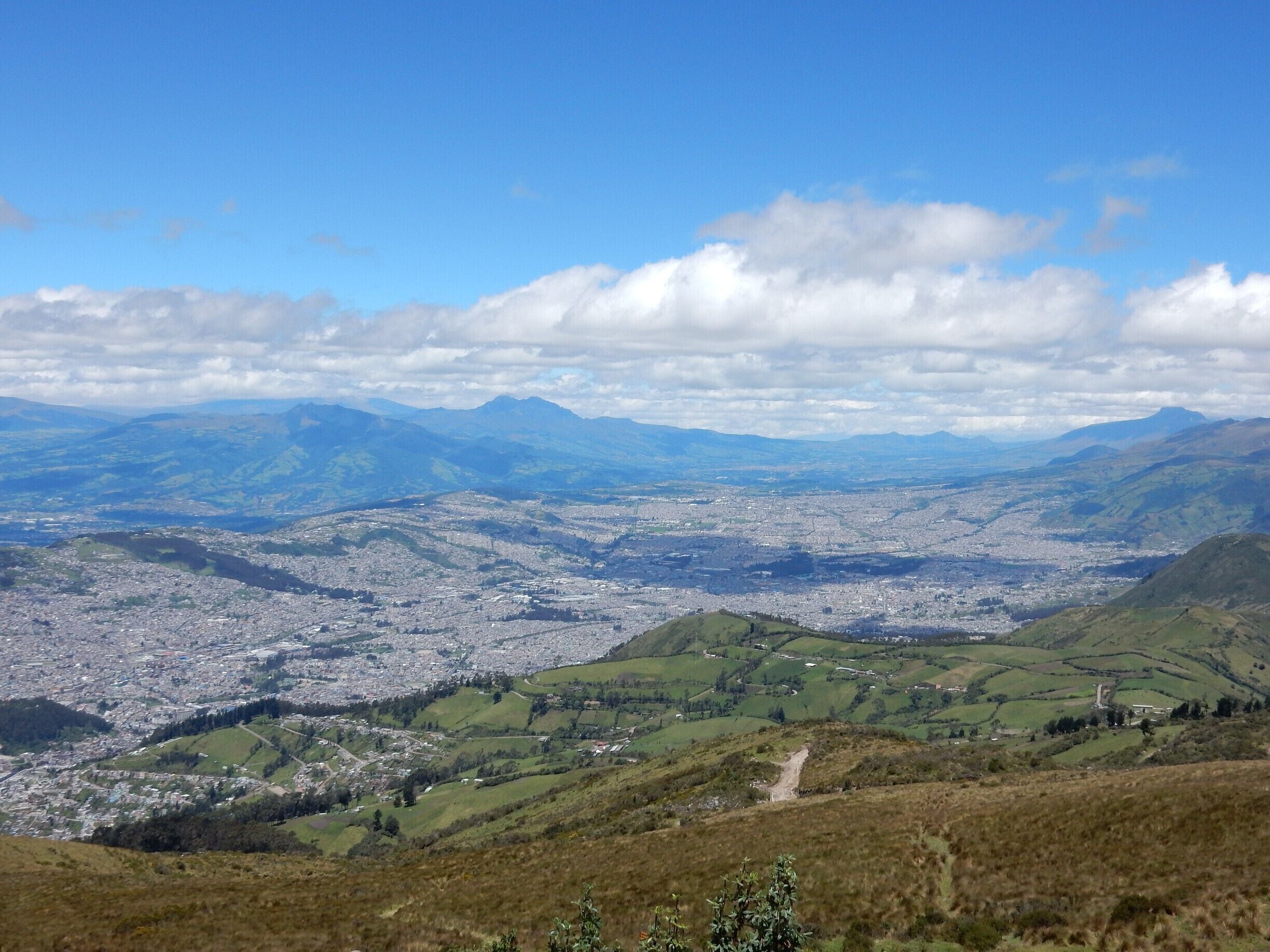 A panoramic view of a city surrounded by rolling hills and distant mountains under a partly cloudy sky.