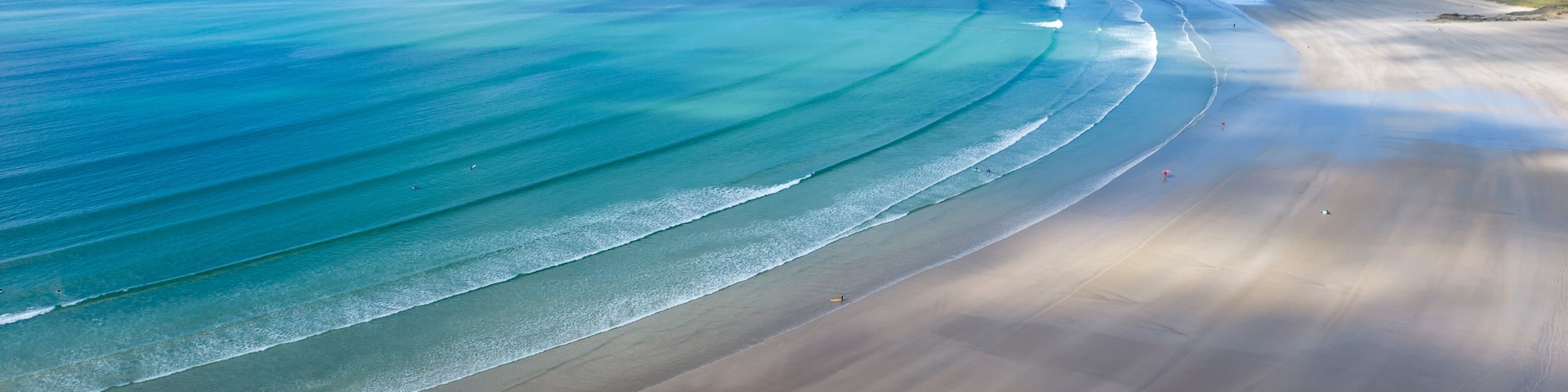 Paisagem costeira com uma extensa praia de areia, ondas suaves e falésias ao longe.