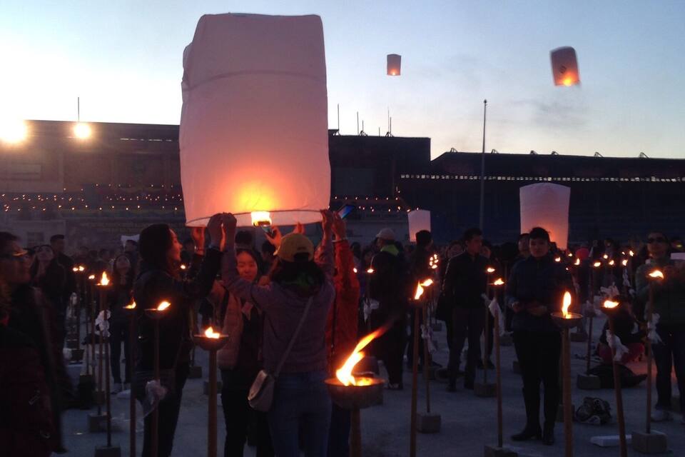 Una multitud de personas en un evento público, con numerosas velas encendidas y farolillos de papel en el cielo.