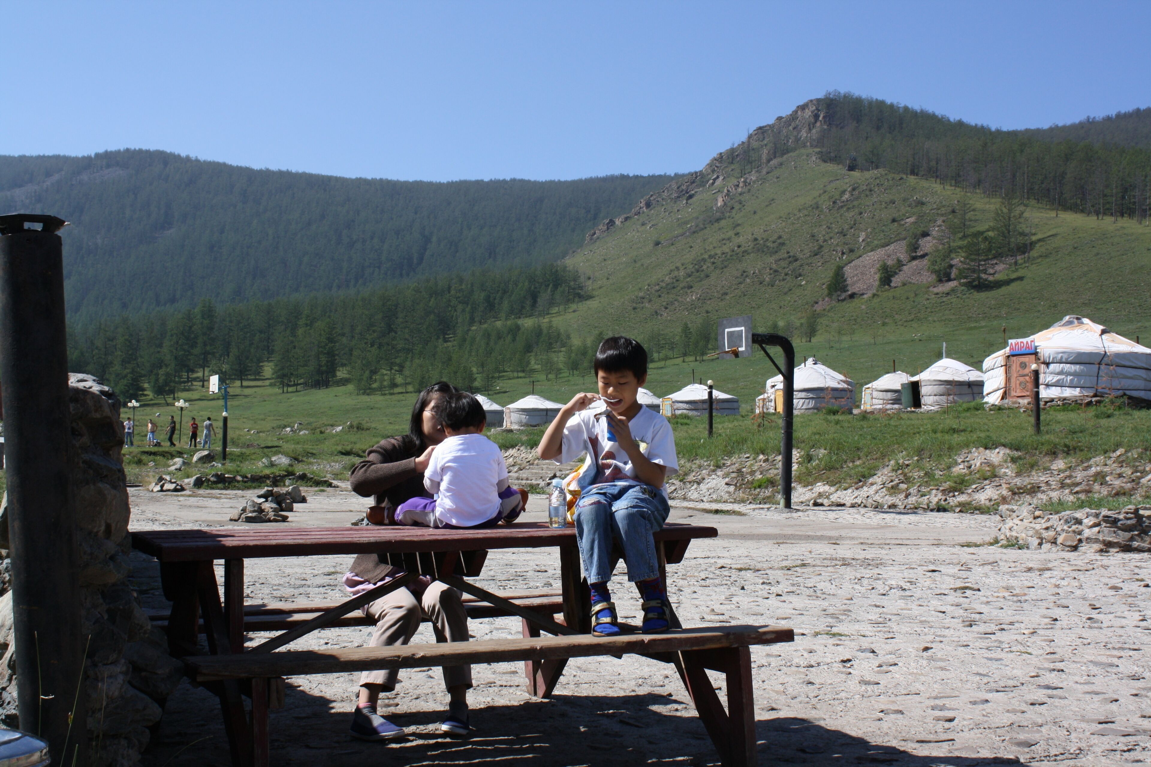 Una mesa de picnic con gente sentada, un paisaje montañoso y yurtas al fondo.