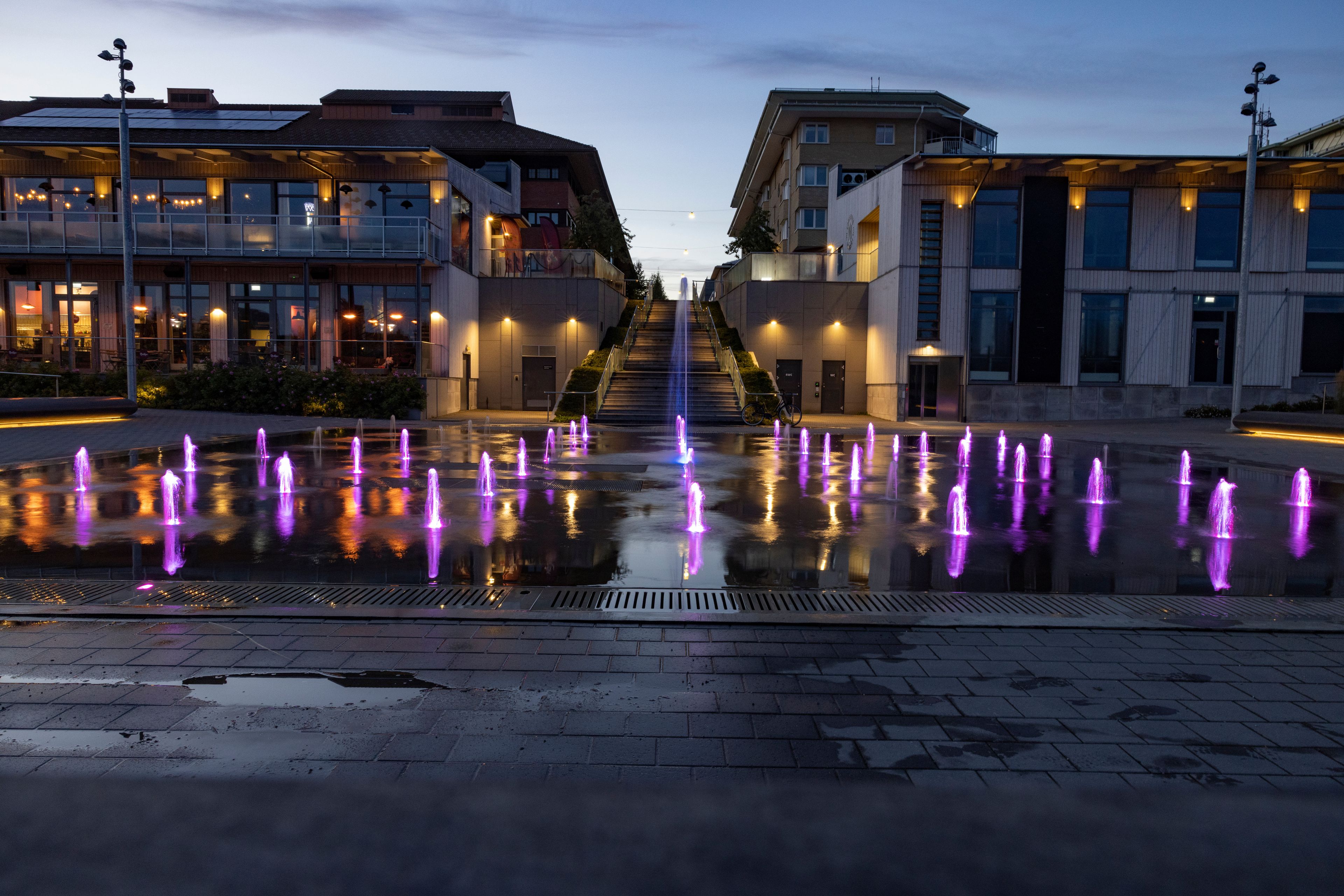 Escena urbana nocturna con un espectáculo de fuentes de agua que reflejan luces, una escalera que conduce a un edificio y arquitectura moderna.