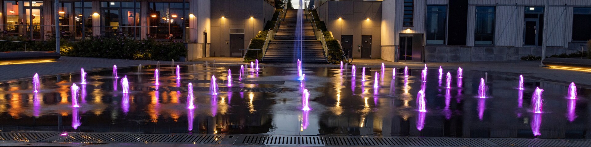 Escena urbana nocturna con un espectáculo de fuentes de agua que reflejan luces, una escalera que conduce a un edificio y arquitectura moderna.