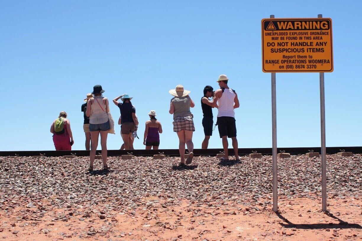 A group of people standing on a rocky terrain with a warning sign about unexploded ordnance.