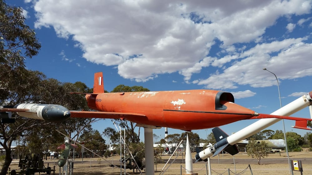 Un avion rouge orné d’une bande blanche sur la queue, exposé à l’extérieur, avec des arbres et un ciel dégagé en arrière-plan.