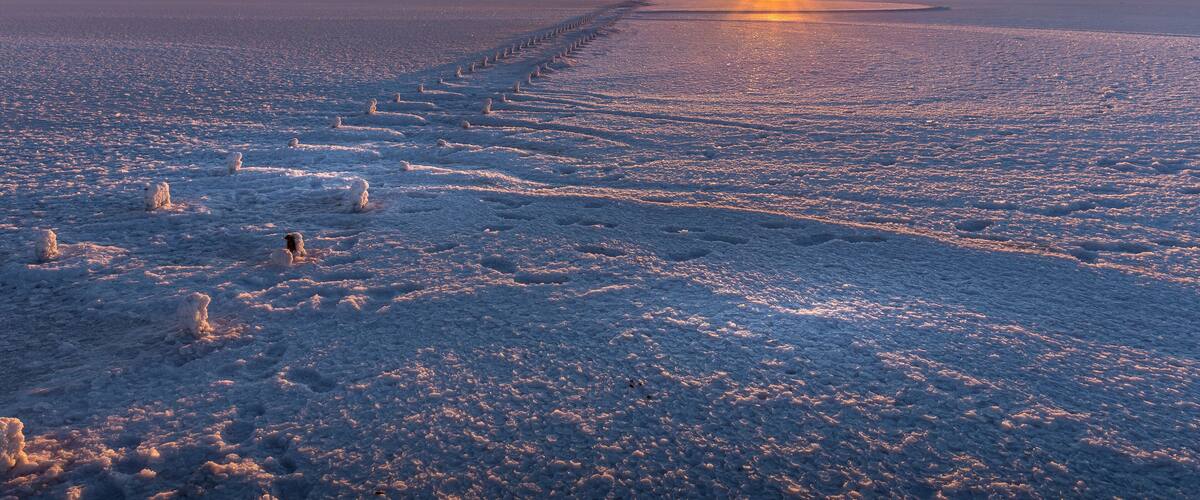 A frozen landscape with a sunset, snow-covered ground, and a clear sky.