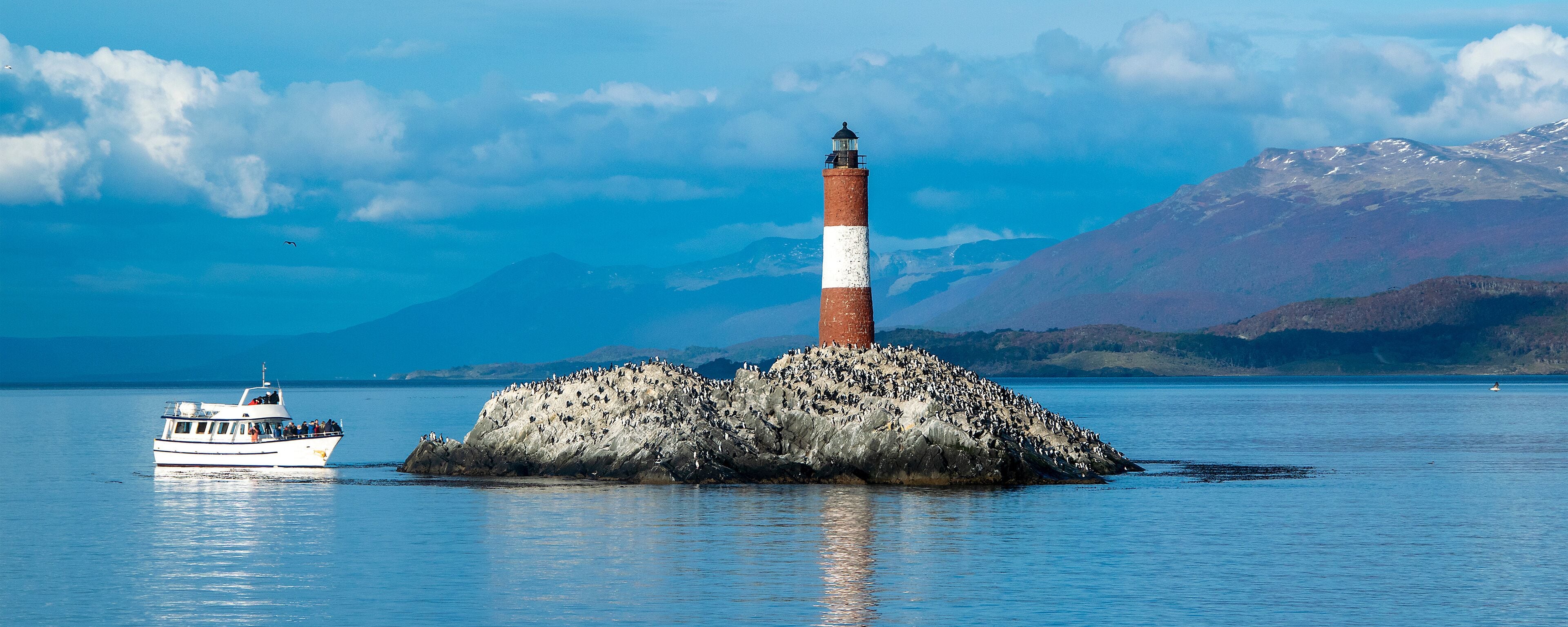 Un faro sobre un promontorio rocoso, con un barco en primer plano y montañas al fondo.