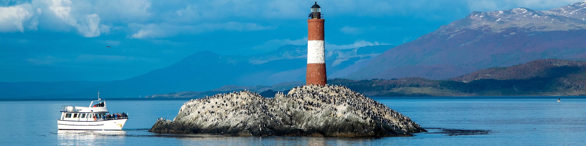 Un faro sobre un promontorio rocoso, con un barco en primer plano y montañas al fondo.