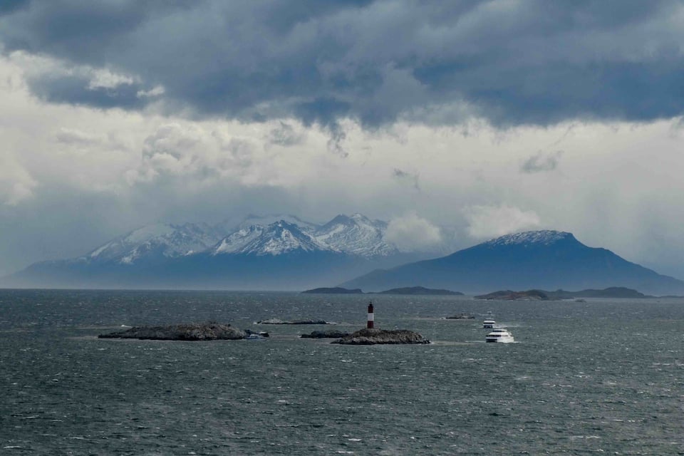 A lighthouse on a rocky outcrop in a body of water with snow-capped mountains in the background.