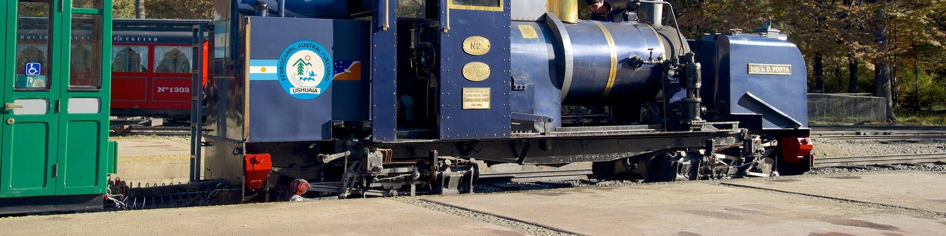 A steam locomotive with a blue and gold logo, a green station building, and trees in the background.