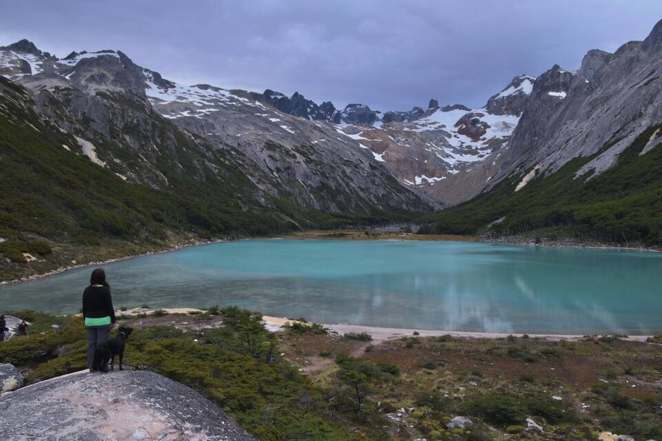 A person standing on a rock overlooking a turquoise lake surrounded by mountains.
