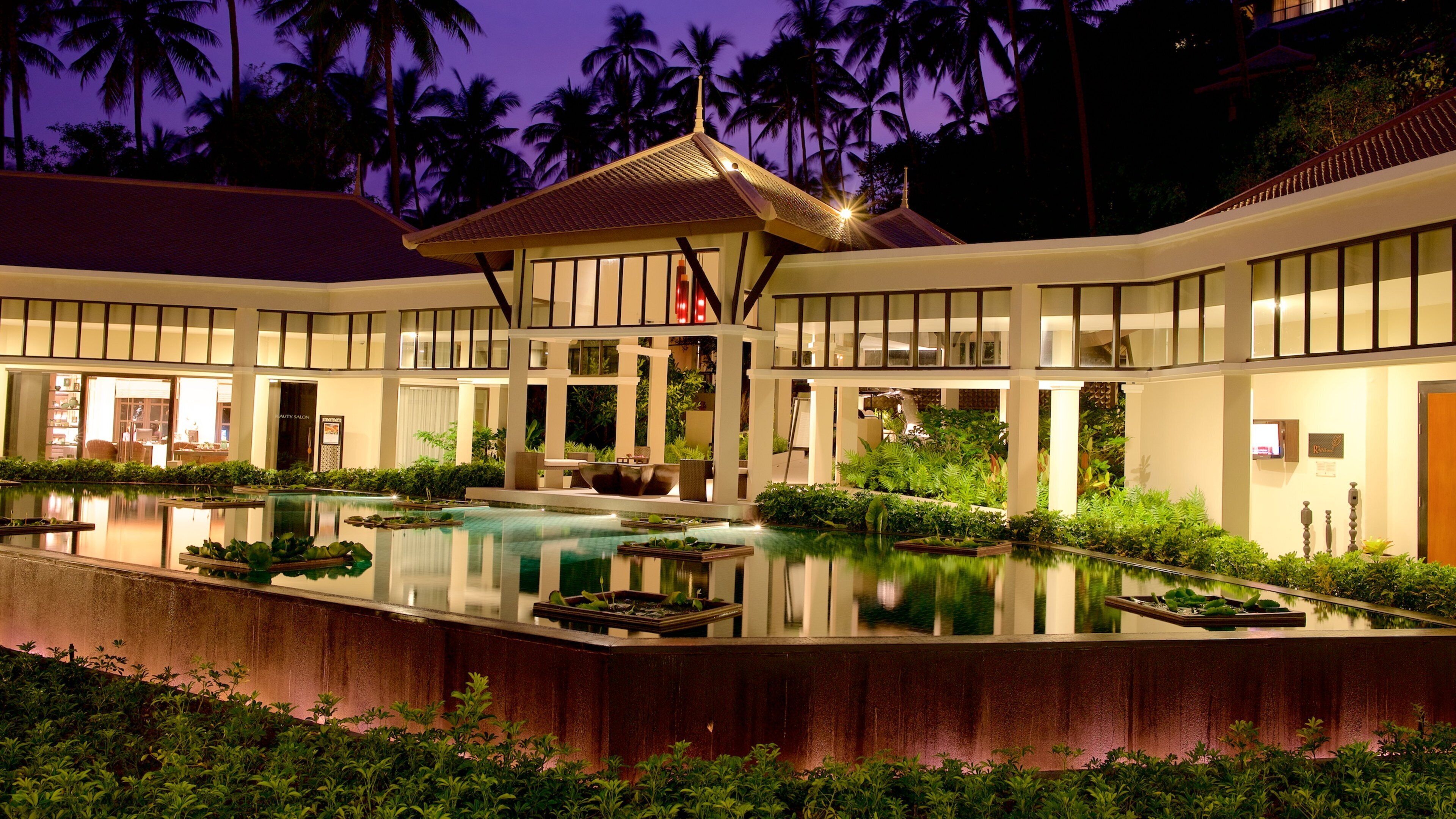 A resort with a swimming pool, palm trees, and a traditional-style building at dusk.