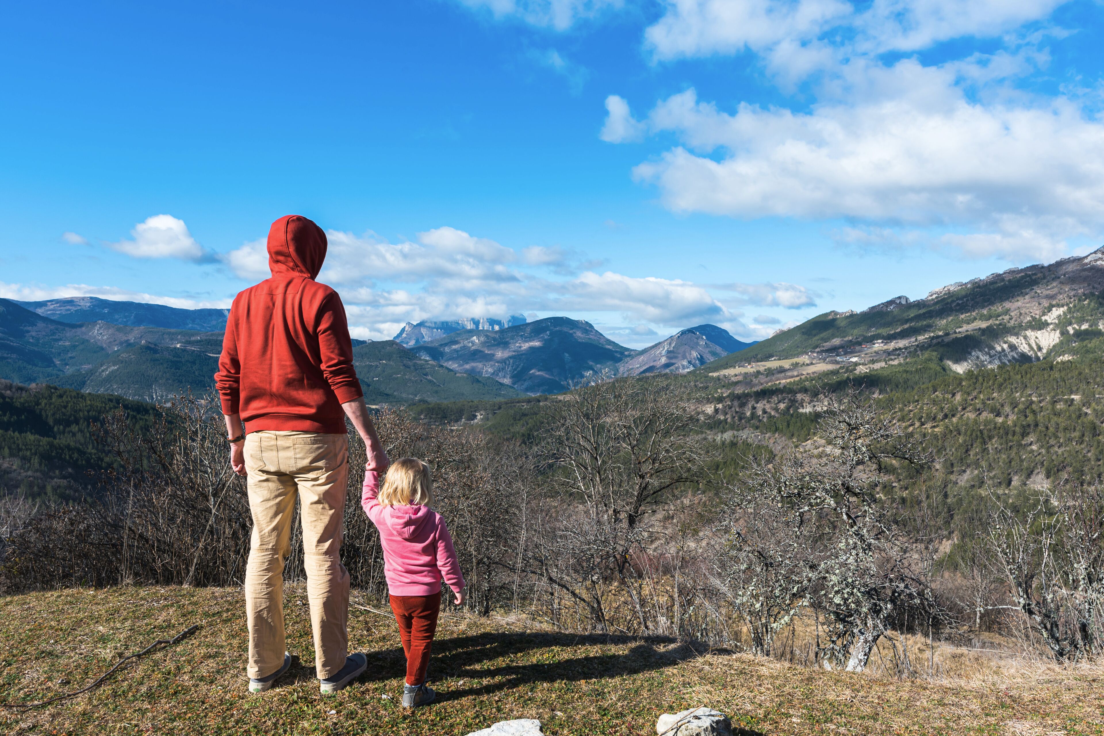 A person in a red hoodie and beige pants, holding hands with a child in a pink jacket, overlooking a mountainous landscape with clear skies.