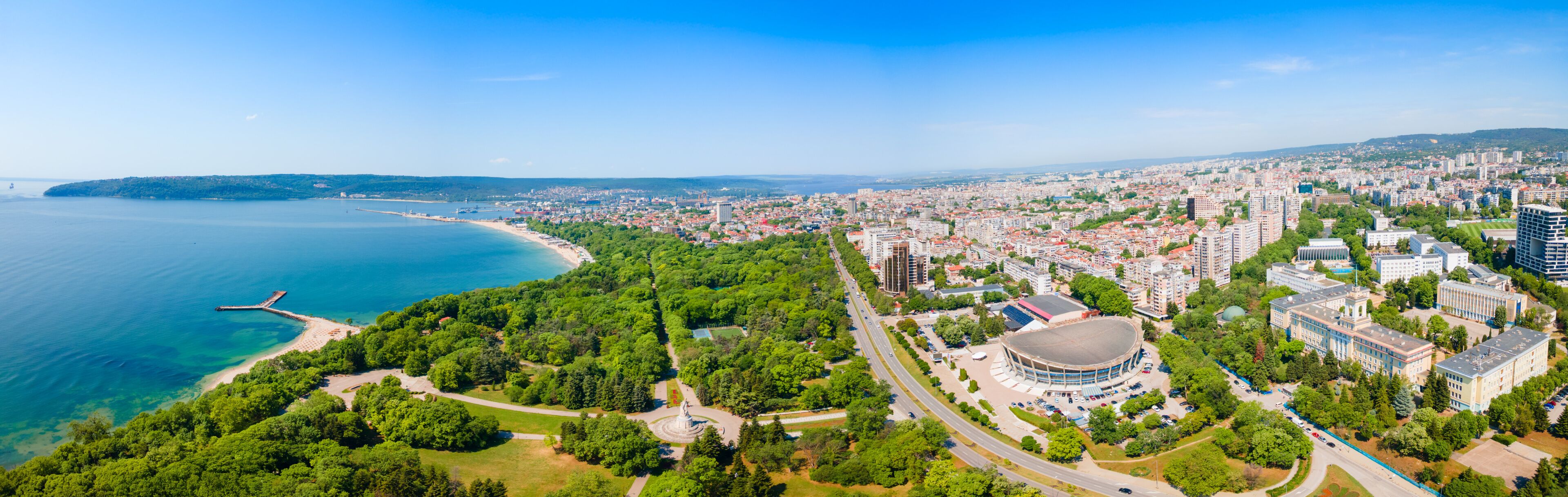 A coastal cityscape with a beach, dense urban area, and a large circular building.
