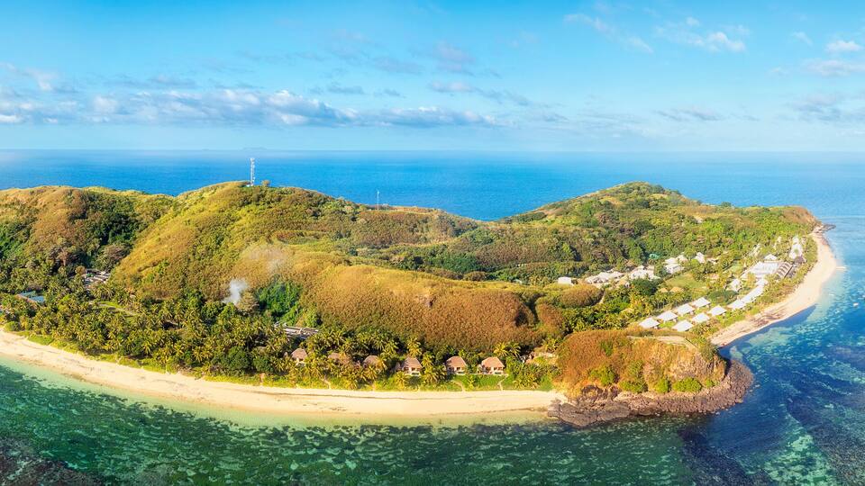 An aerial view of a tropical island with a crescent-shaped coastline, clear turquoise waters, and lush greenery.