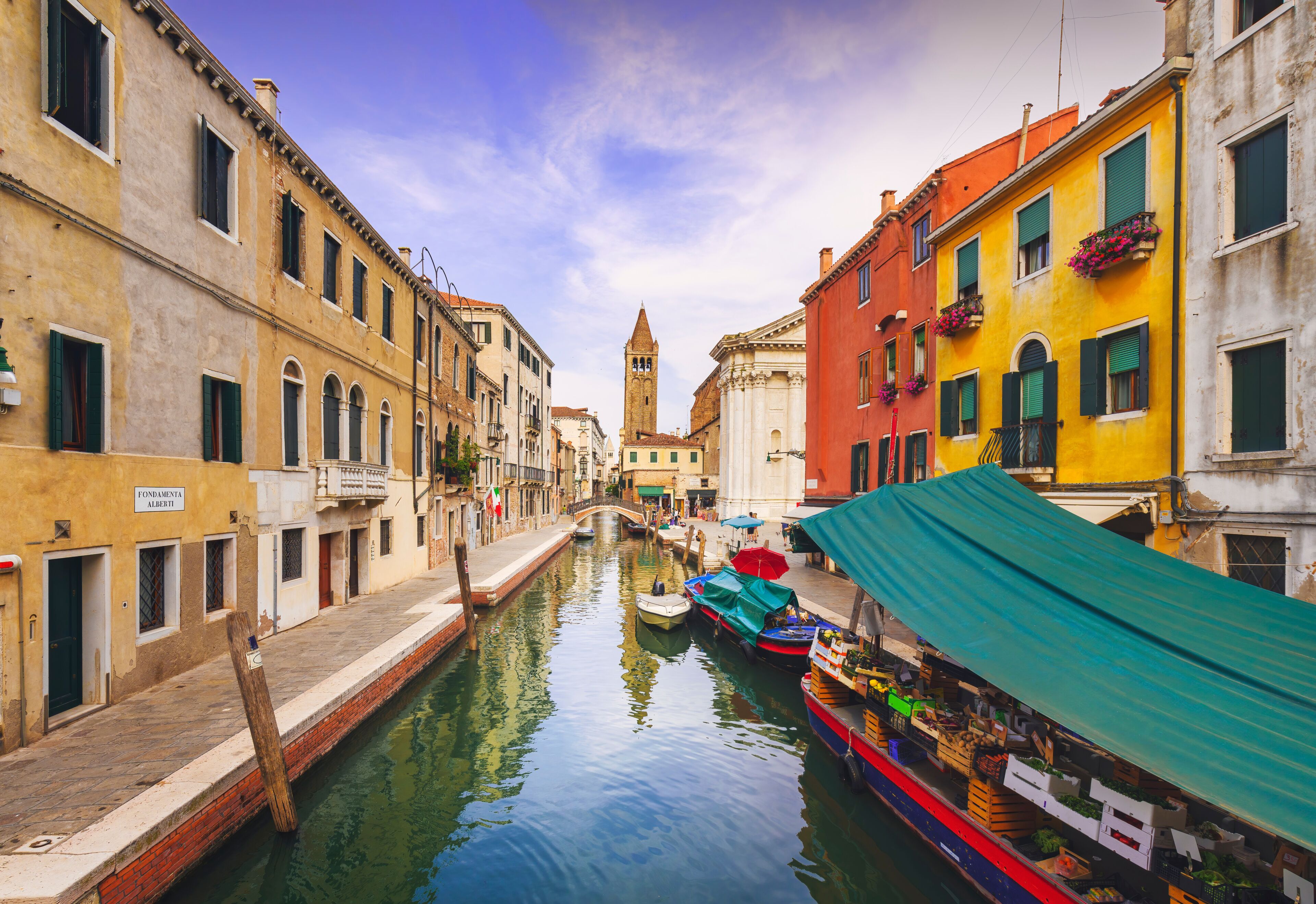Un estrecho canal veneciano bordeado por edificios coloridos, un puente y una campanario.