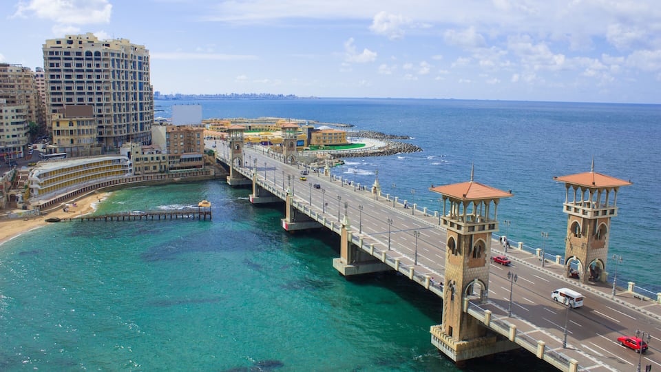 A coastal bridge with clear turquoise waters, a beach, and urban buildings.