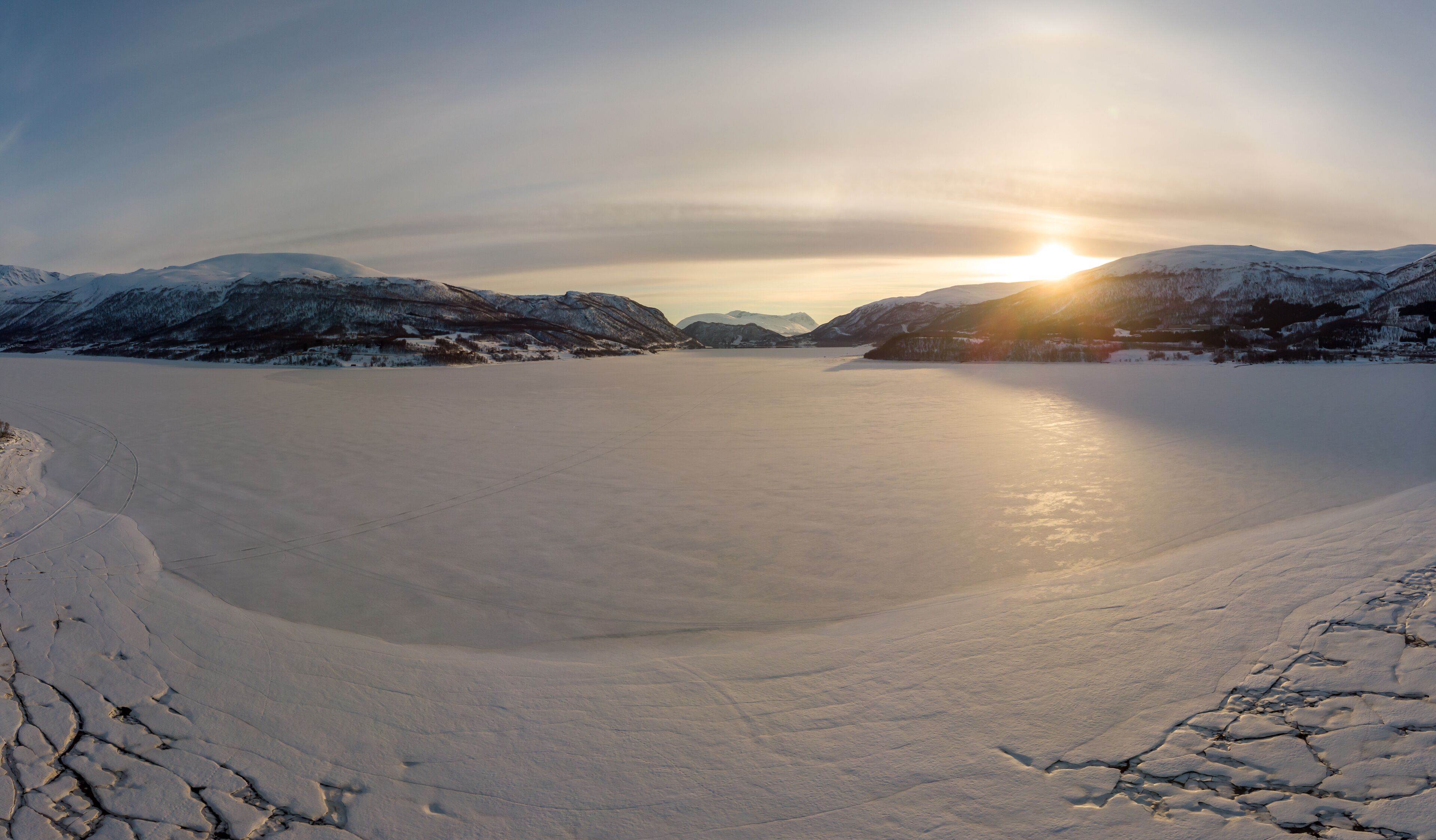 A frozen lake surrounded by snow-covered mountains during sunset.