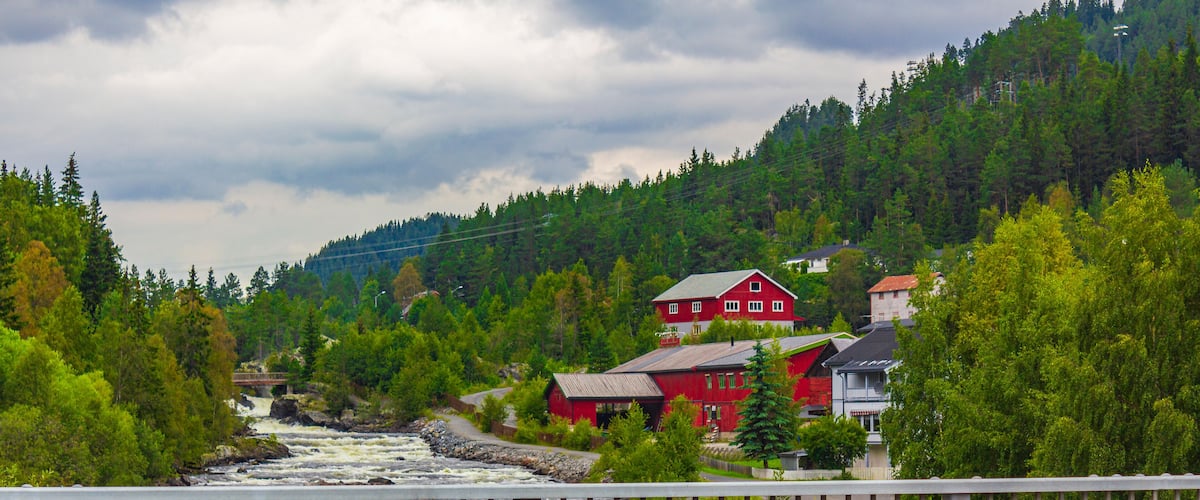 En flod der løber gennem en skovdal med røde bygninger og en bro.