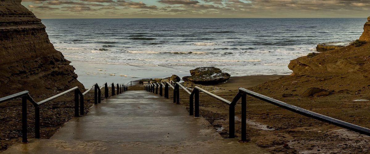 A coastal walkway with metal railings leading to the sea, flanked by rocky cliffs.