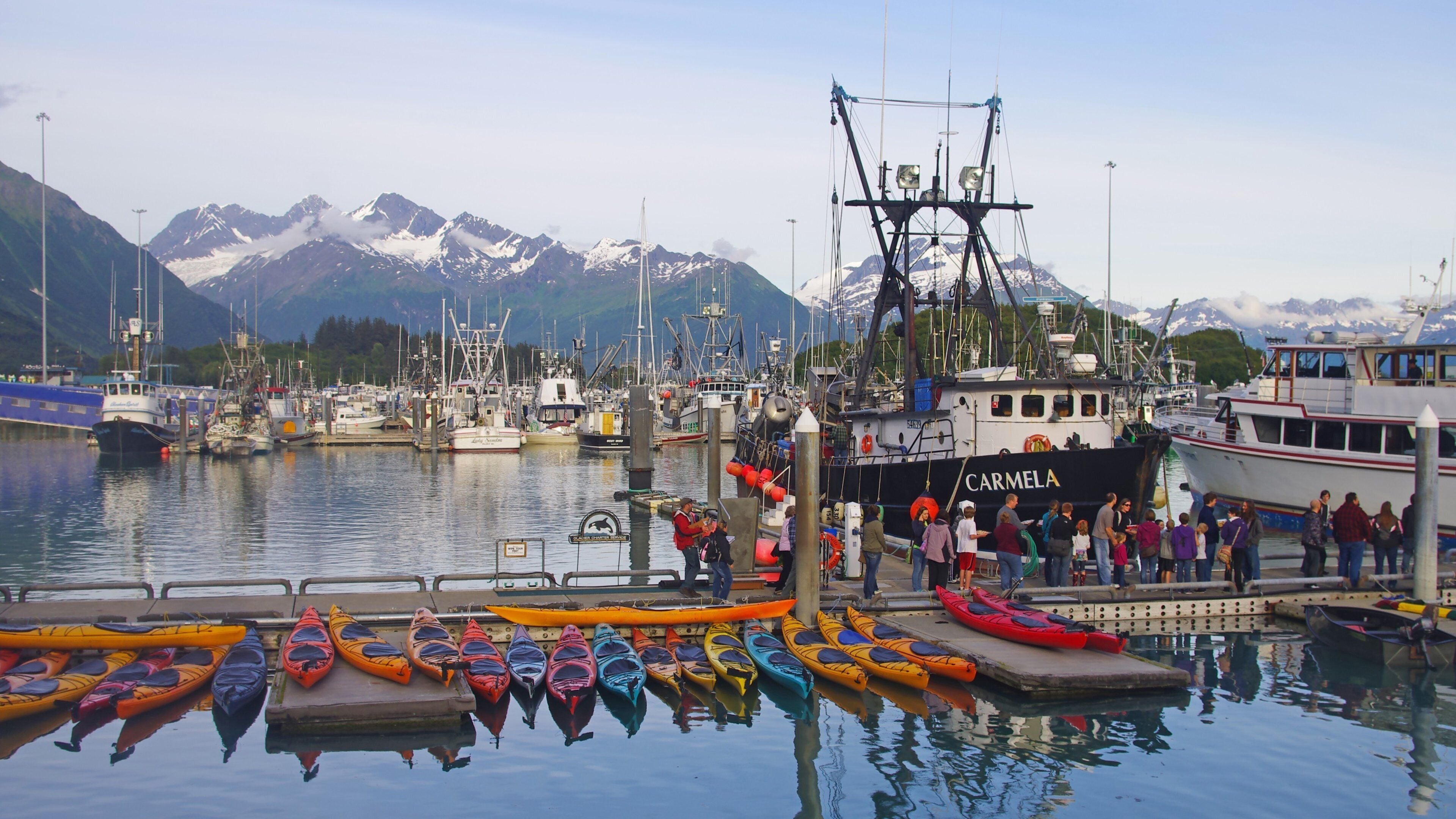 A harbor with boats, a large ship named 'CARMELA', and mountains in the background.