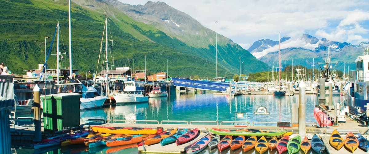A harbor with colorful kayaks, boats, and a mountain backdrop.