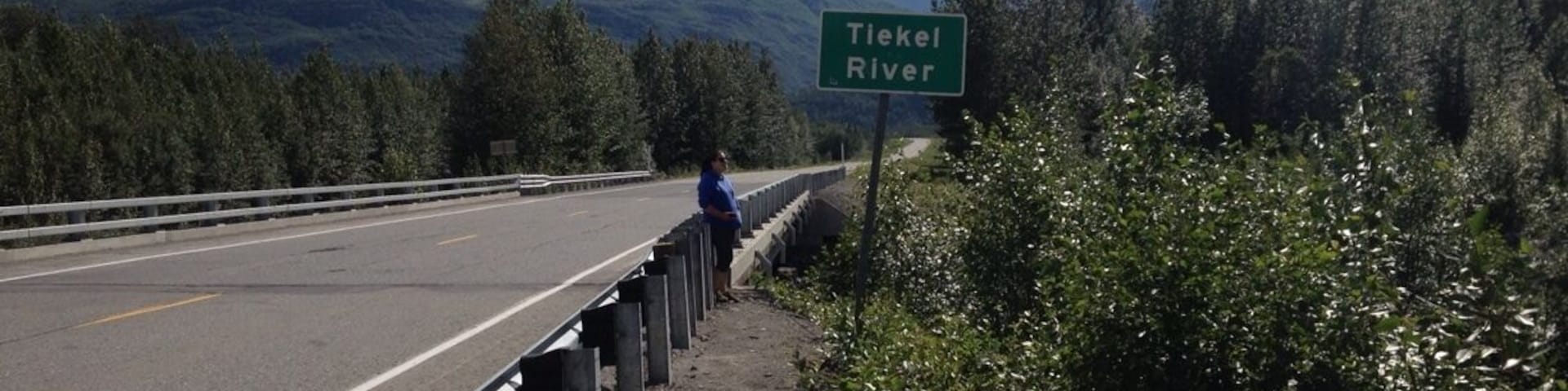 Uma estrada com guarda-corpo, uma pessoa ao lado do guarda-corpo e uma placa indicando Rio Tiekel.