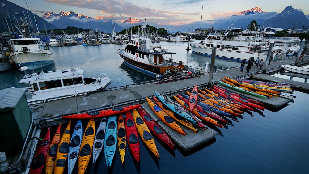 Un port de plaisance où sont amarrés des bateaux et des kayaks aux couleurs vives, le tout entouré de montagnes.