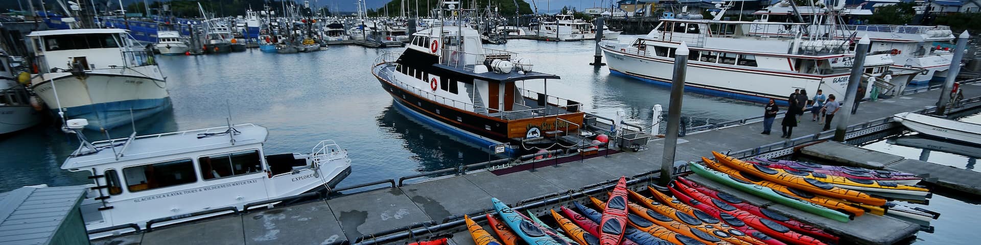 A marina with boats and colorful kayaks docked, surrounded by mountains.