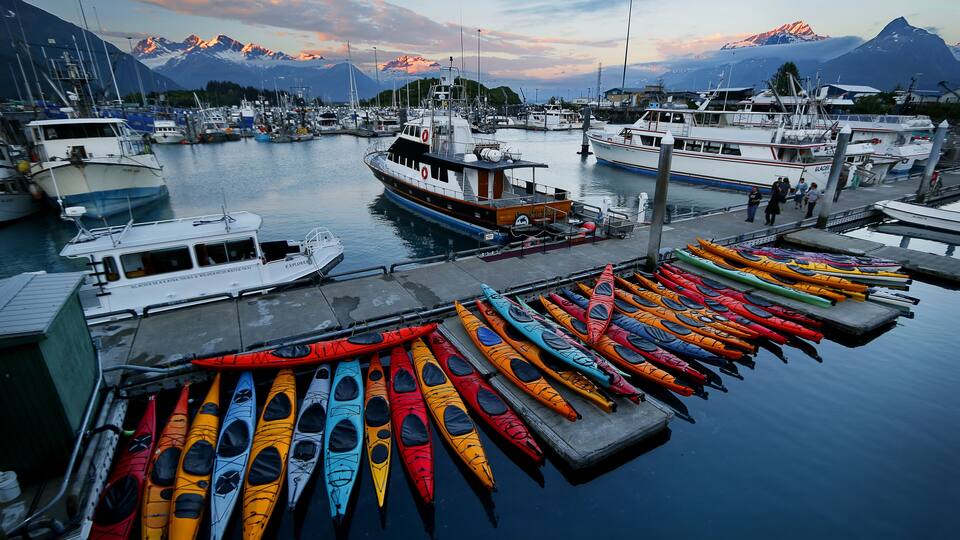A marina with boats and colorful kayaks docked, surrounded by mountains.