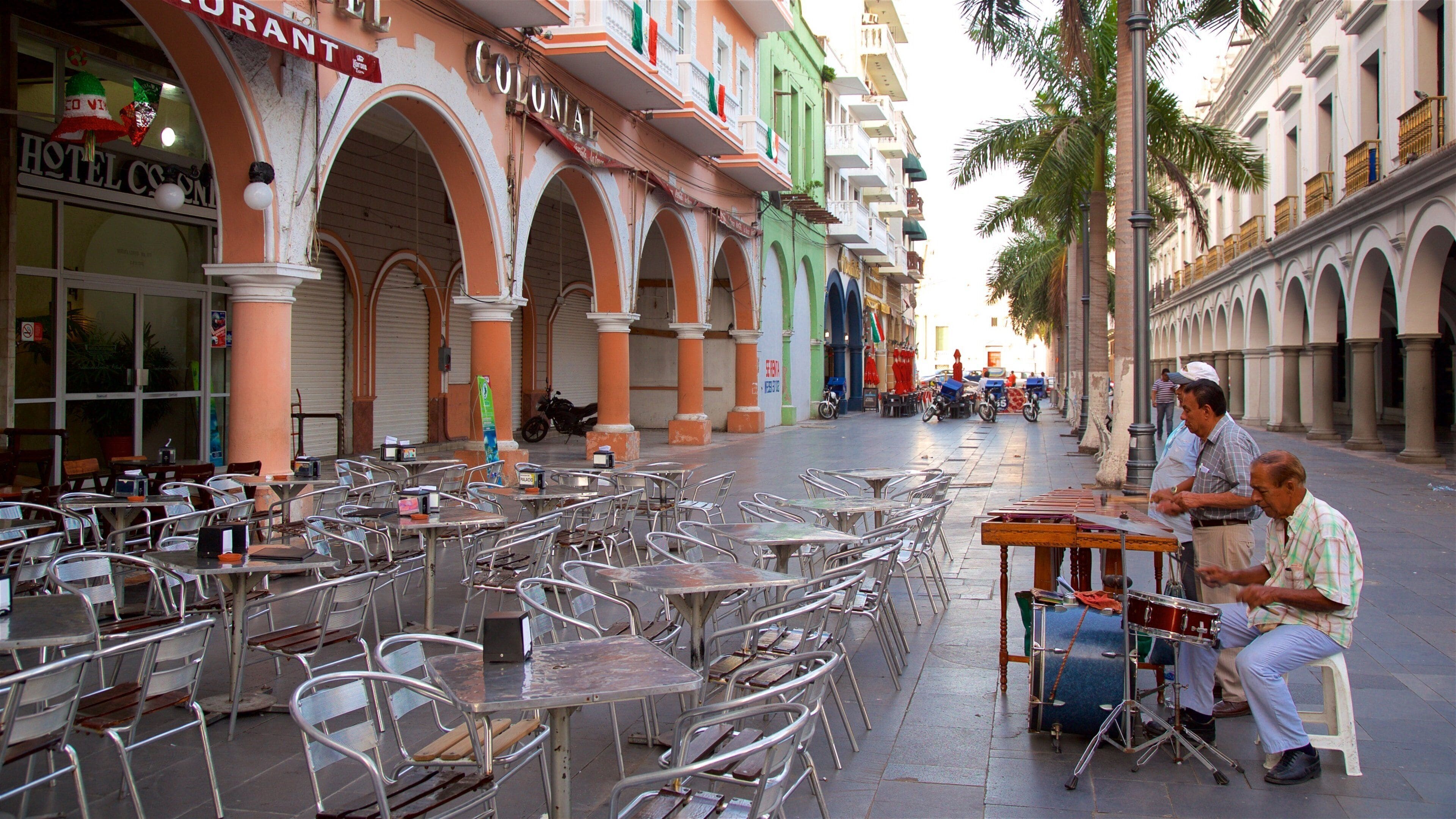 Outdoor cafe seating with tables and chairs, a street with buildings, and a musician playing a drum set.