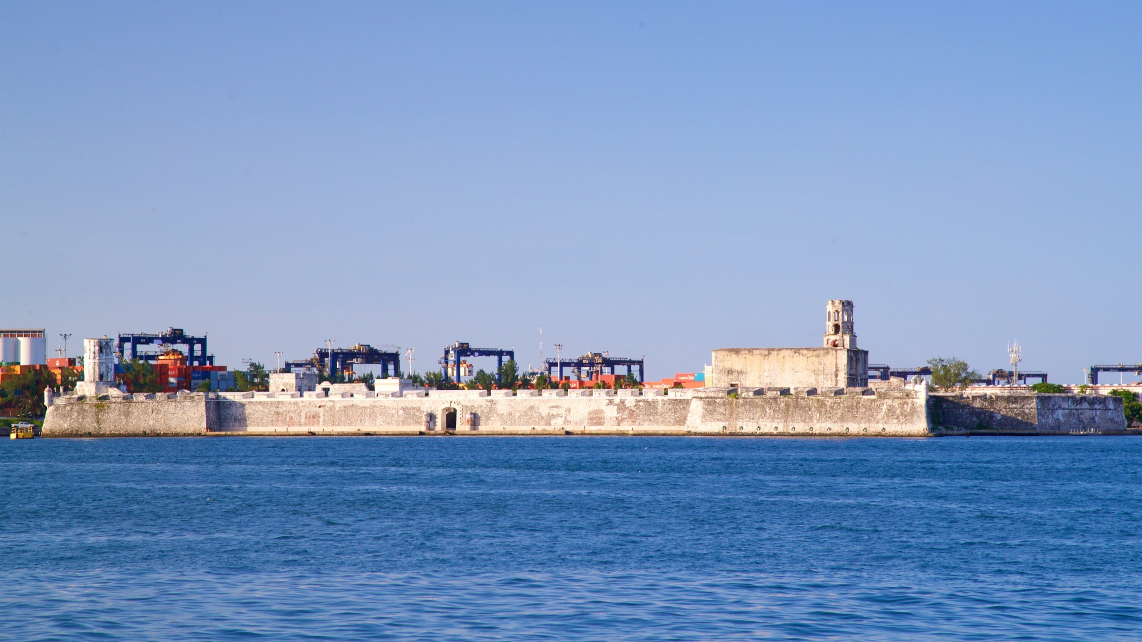 A coastal cityscape with a historic fort, a prominent tower, and industrial cranes.