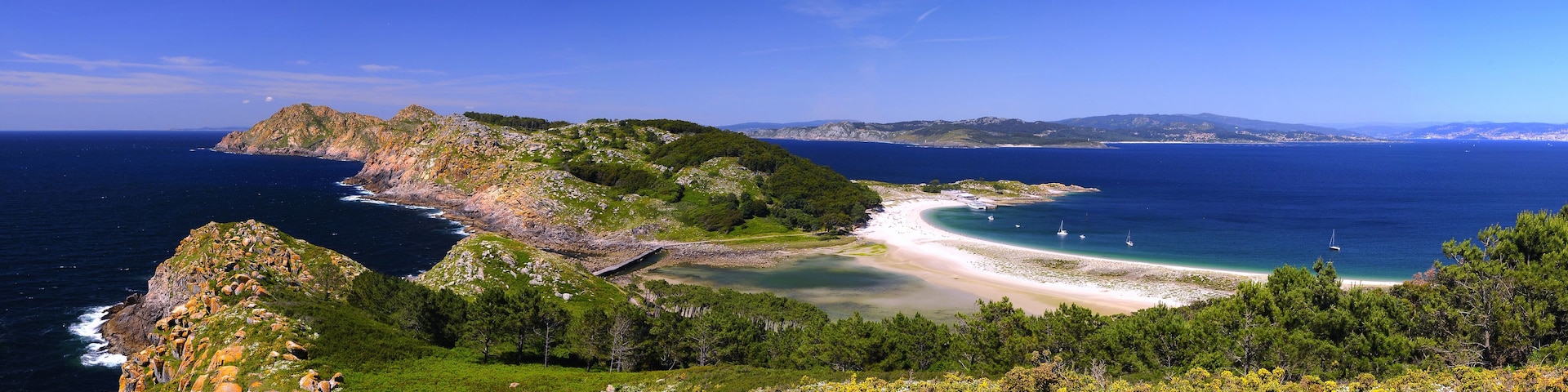 Un paisaje costero con una playa, un afloramiento rocoso y un cielo azul despejado.