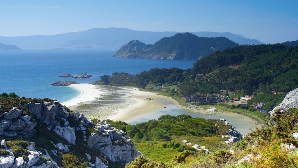 A coastal landscape with a sandy beach, rocky outcrops, and dense forested hills.