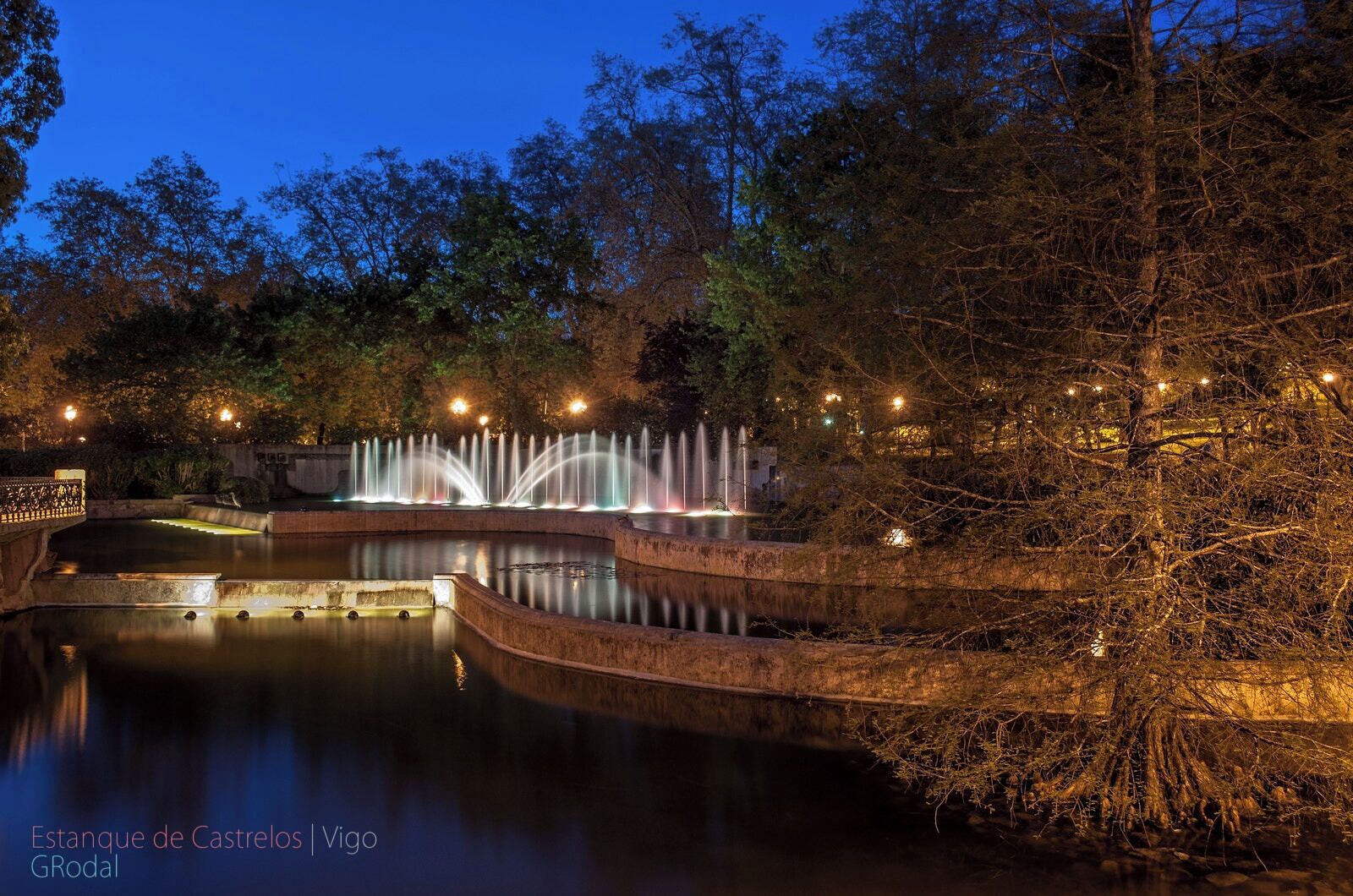 A nighttime view of the Estanque de Castrelos with illuminated fountains and a bridge.