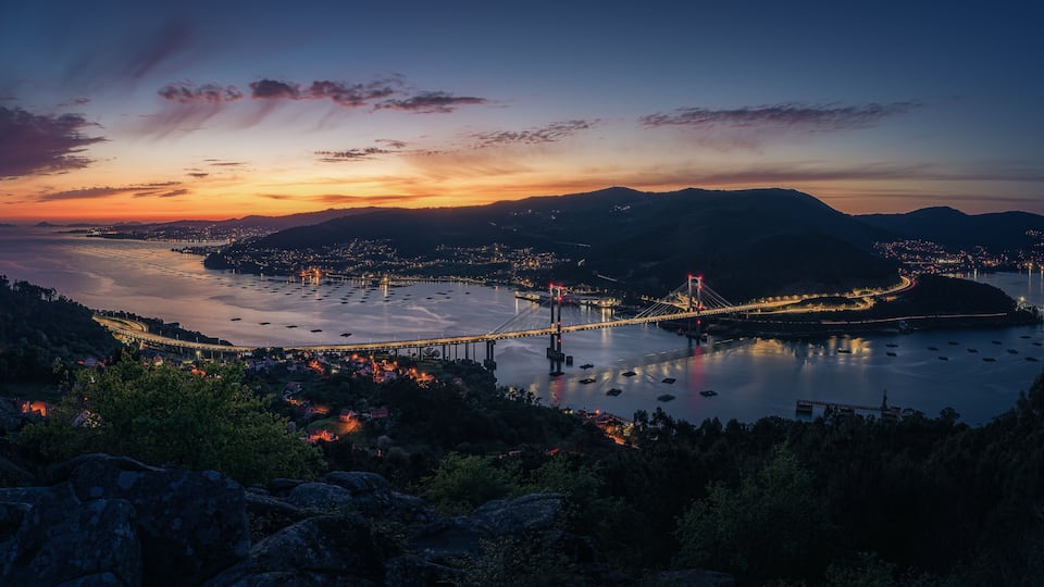 A coastal cityscape at dusk with a prominent bridge, calm water, and surrounding hills.