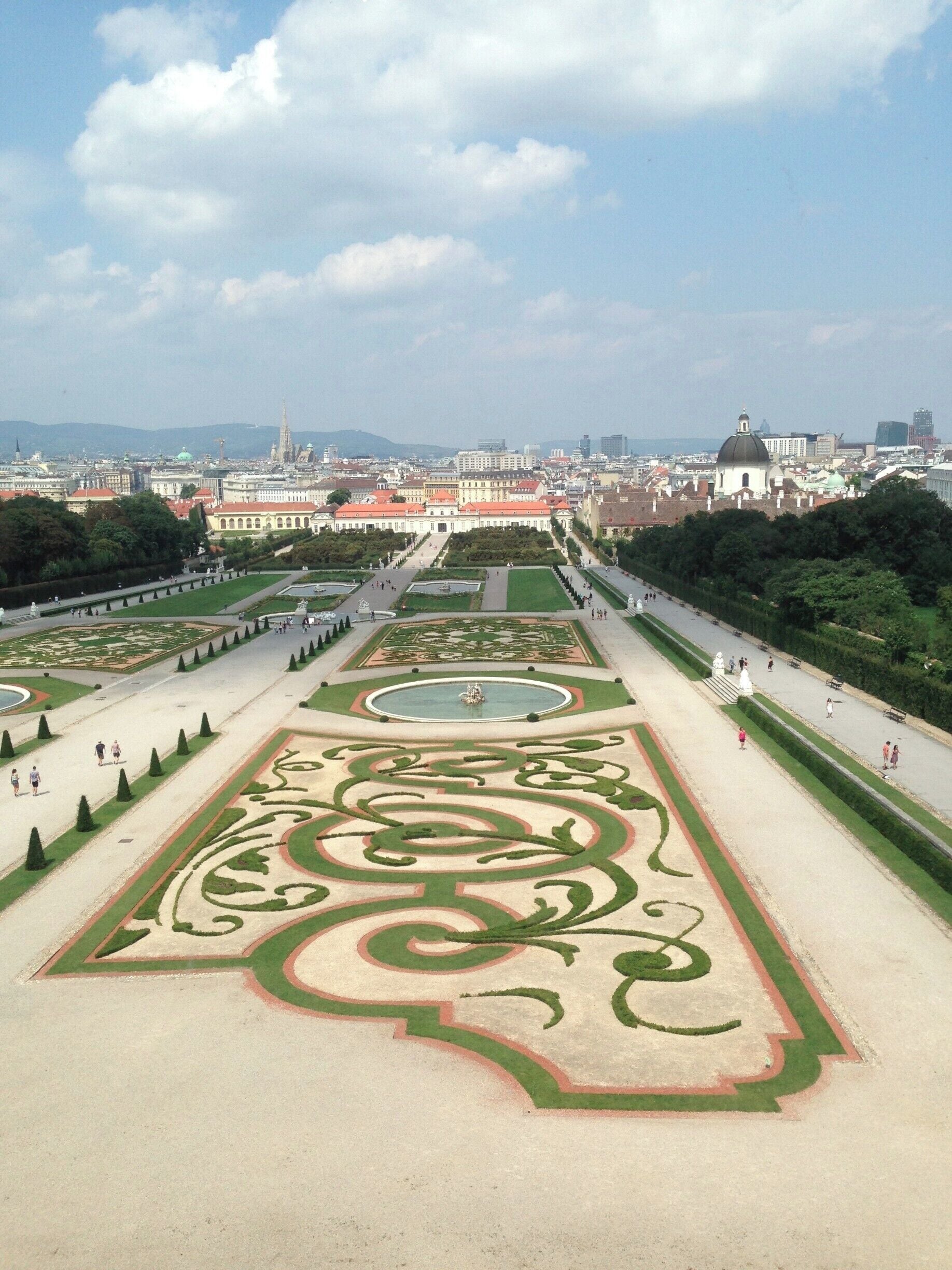 Ein prächtiger, formeller Garten mit geometrischen Mustern, einer zentralen Fontäne und einem entfernten Blick auf eine Stadtlandschaft mit historischen Gebäuden.