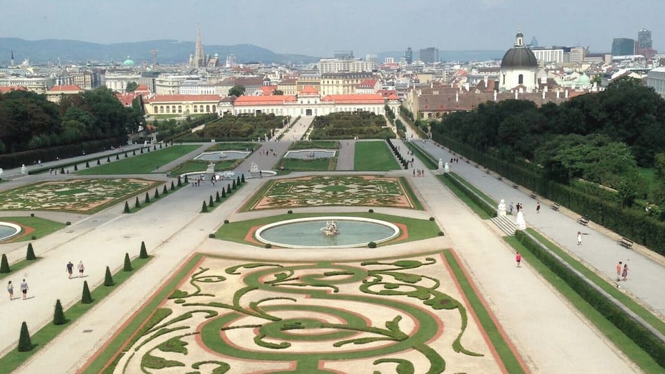 Ein prächtiger, formeller Garten mit geometrischen Mustern, einer zentralen Fontäne und einem entfernten Blick auf eine Stadtlandschaft mit historischen Gebäuden.