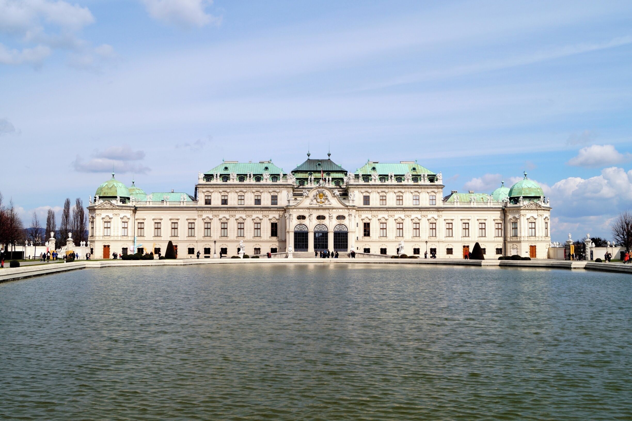 Un grand bâtiment imposant, orné de coupoles vertes et doté d’une façade symétrique, entouré d’une étendue d’eau.