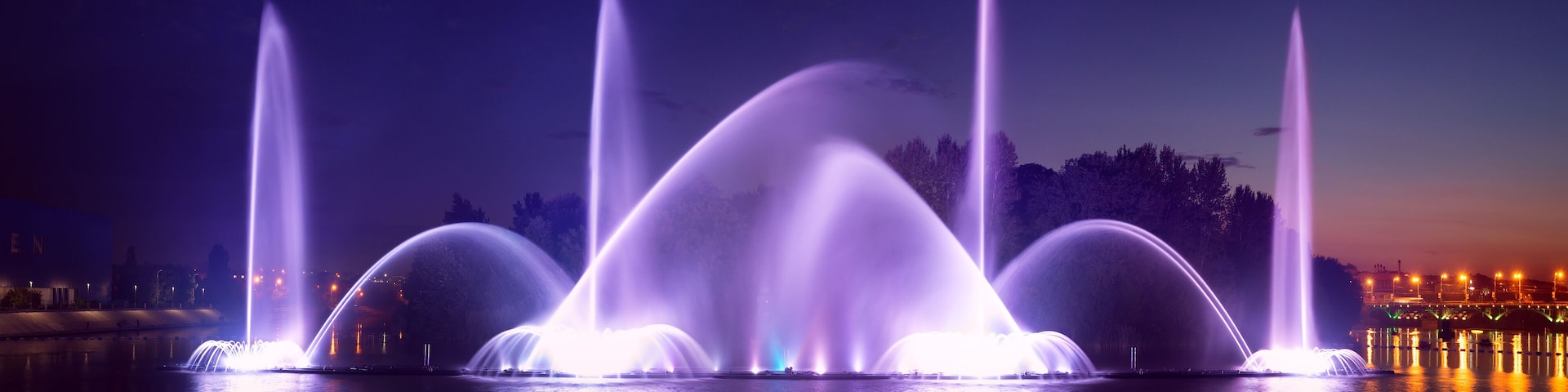 A nighttime fountain display with multiple water jets illuminated in purple.