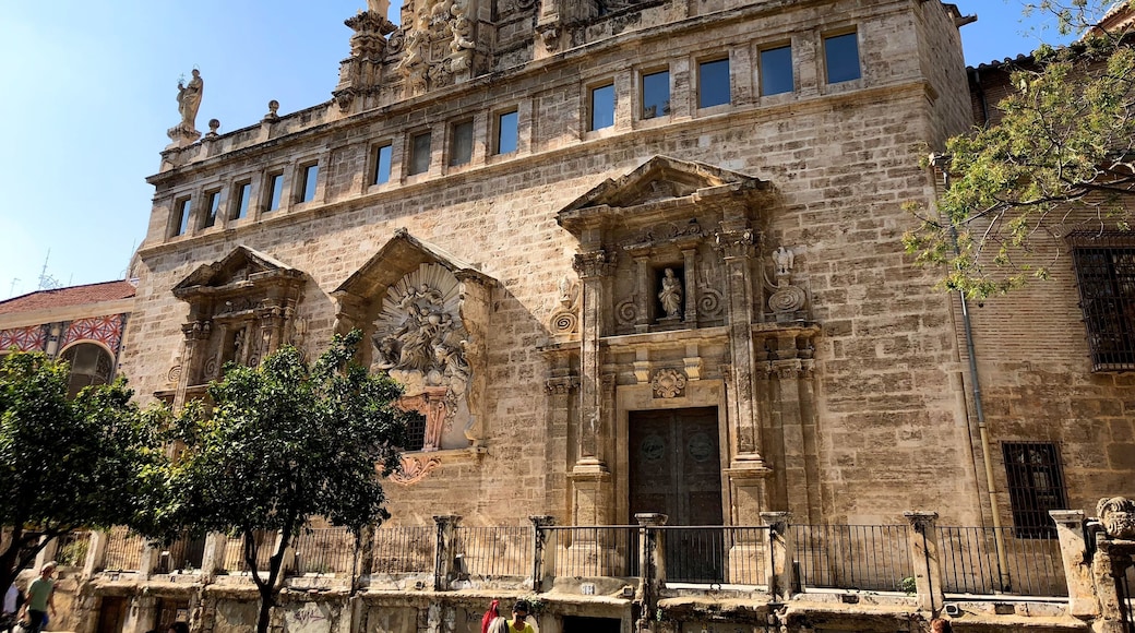 The primitive church of Sant Joan Mercat was erected in the suburb of the city known as the Boatella. From its old Gothic structure there is still the nave and the large blind oculus, known as "La O de Sant Joan", which was conceived as a large rosette on the façade of the feet.
In 1592 the church suffered a spectacular fire that forced an almost total reconstruction throughout the seventeenth and eighteenth centuries. It stands out for the infrequent the wide façade of the Market, conceived as a grandiose stone altarpiece on a terrace that dominates the square in front of the Lonja, forming a unique urban set.
It is presided by the sculpture of the Virgin of the Rosary, work in stucco by Jacopo Bertesi, and on it stands the clock tower, flanked by the two Santos Juanes and crowned by the famous "Pardal de Sant Joan", the weather vane to which -according to tradition- was made to look at the children when their humble parents abandoned them in the square. In the lower part of the terrace there are "les covetes de Sant Joan", semi-basements in which once were junkyards and old shops, part of the living history of Valencia.
The interior of the temple is an overflowing Baroque imagination, with statues of Bertesi representing characters of the Twelve Tribes of Israel, and especially the set of frescoes that Antonio Palomino executed in the vault.