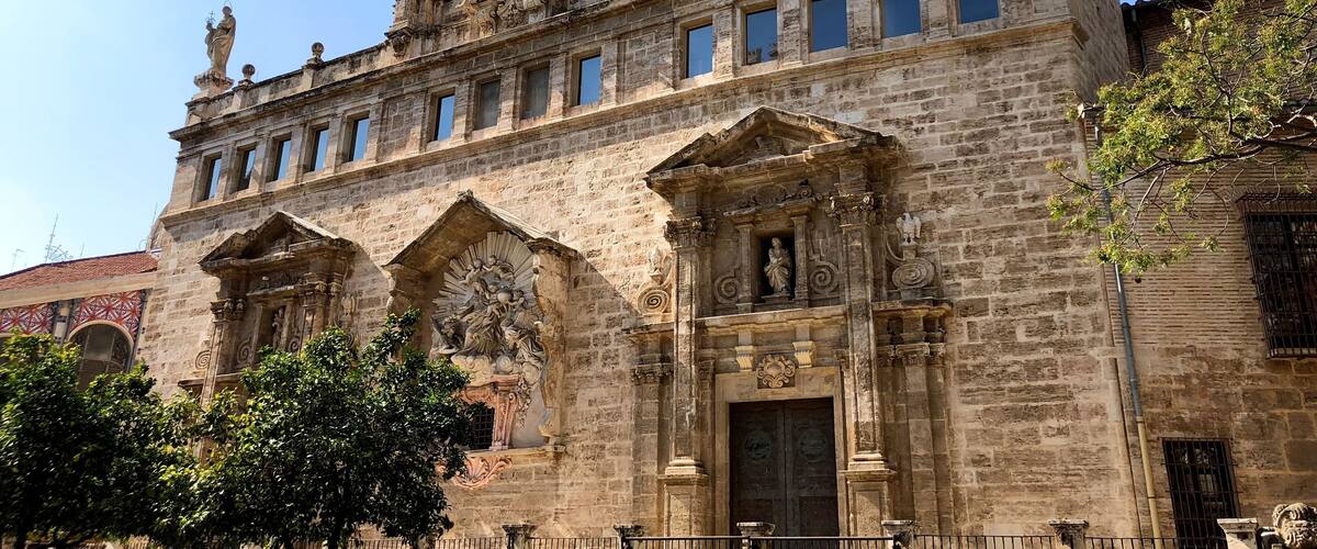 The primitive church of Sant Joan Mercat was erected in the suburb of the city known as the Boatella. From its old Gothic structure there is still the nave and the large blind oculus, known as "La O de Sant Joan", which was conceived as a large rosette on the façade of the feet.
In 1592 the church suffered a spectacular fire that forced an almost total reconstruction throughout the seventeenth and eighteenth centuries. It stands out for the infrequent the wide façade of the Market, conceived as a grandiose stone altarpiece on a terrace that dominates the square in front of the Lonja, forming a unique urban set.
It is presided by the sculpture of the Virgin of the Rosary, work in stucco by Jacopo Bertesi, and on it stands the clock tower, flanked by the two Santos Juanes and crowned by the famous "Pardal de Sant Joan", the weather vane to which -according to tradition- was made to look at the children when their humble parents abandoned them in the square. In the lower part of the terrace there are "les covetes de Sant Joan", semi-basements in which once were junkyards and old shops, part of the living history of Valencia.
The interior of the temple is an overflowing Baroque imagination, with statues of Bertesi representing characters of the Twelve Tribes of Israel, and especially the set of frescoes that Antonio Palomino executed in the vault.