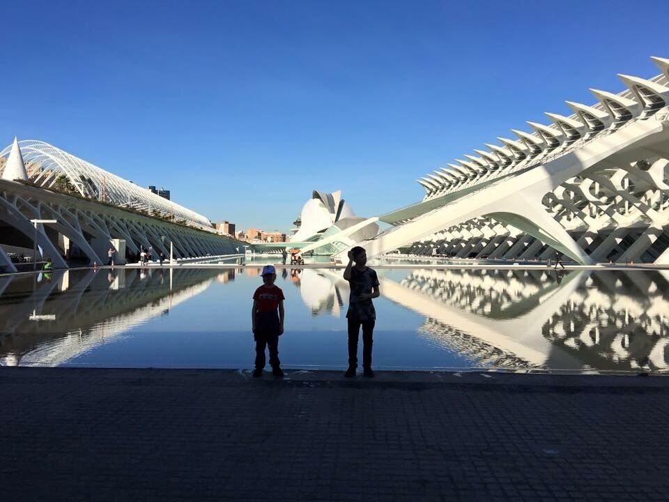 Twee personen staan bij een spiegelglad wateroppervlak met moderne architectuur op de achtergrond.