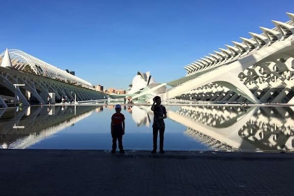 Two people standing by a reflective water surface with modern architectural structures in the background.