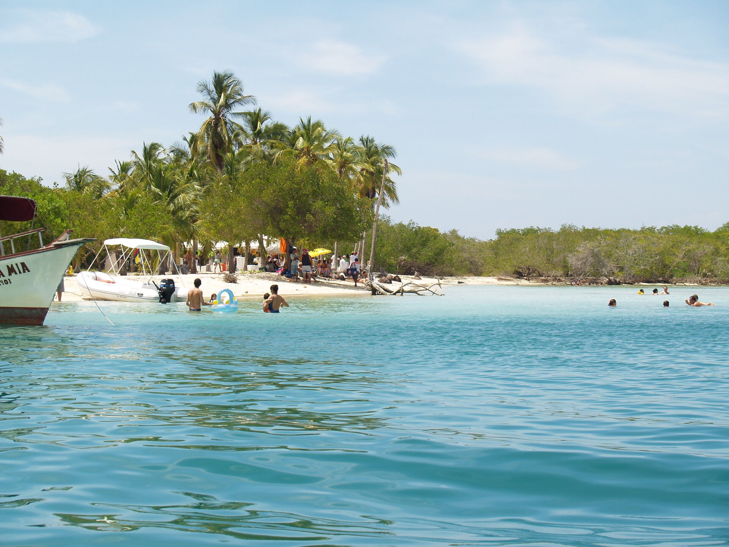 A beach with palm trees, people swimming, and boats in the water.