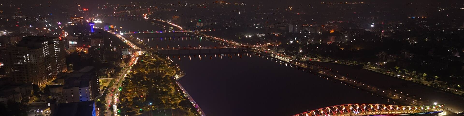 A cityscape at night with a brightly lit bridge spanning a river, surrounded by illuminated buildings and streets.