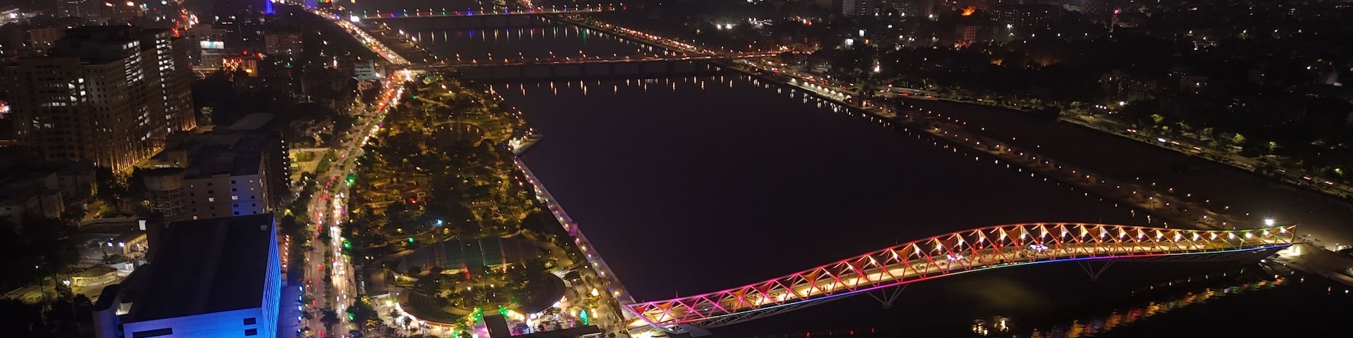 Nachtansicht einer Stadt mit einer hell beleuchteten Brücke über einem Fluss, umgeben von beleuchteten Gebäuden und Straßen.