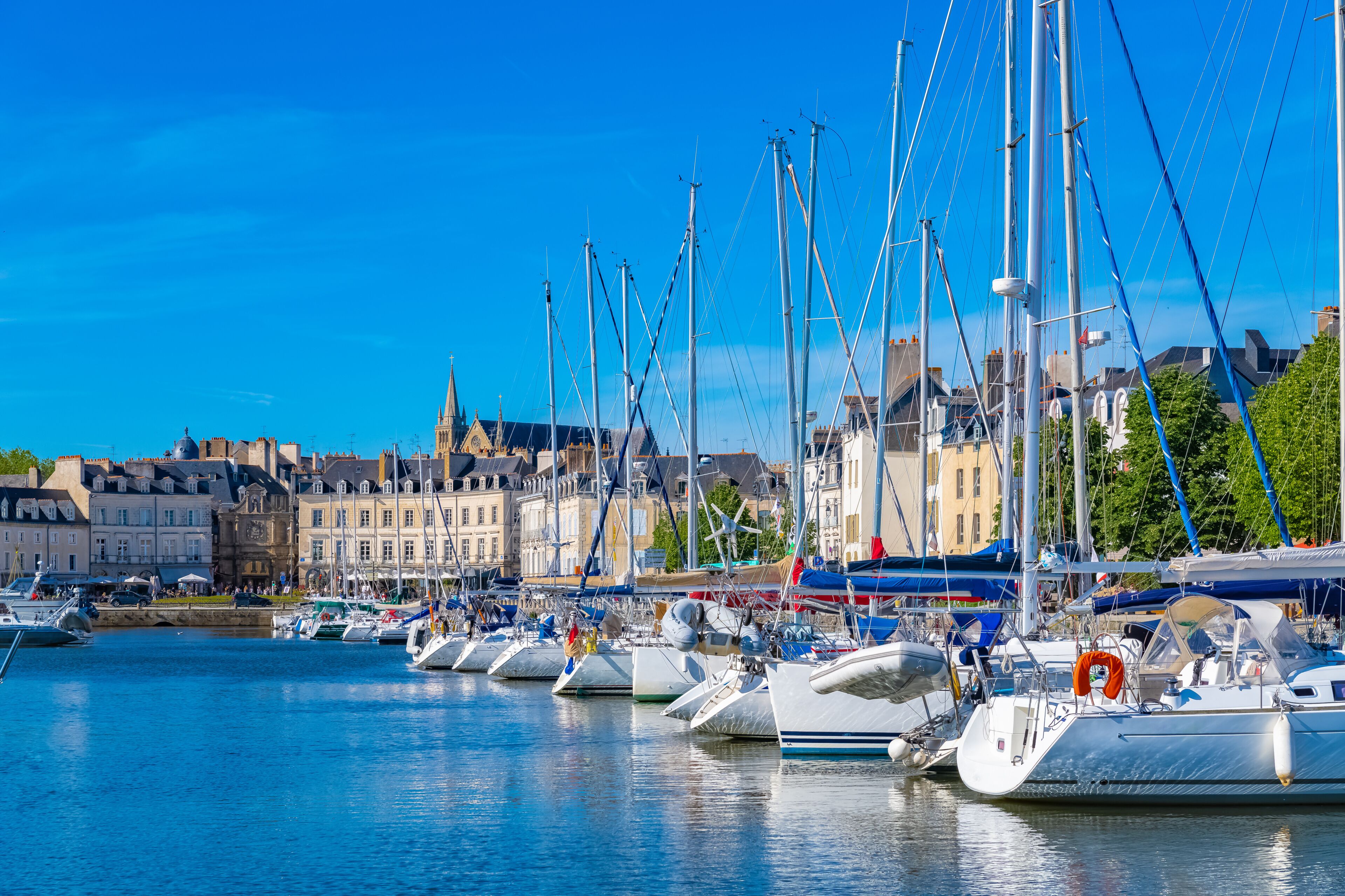 A marina with multiple sailboats docked, a clear blue sky, and historic buildings in the background.
