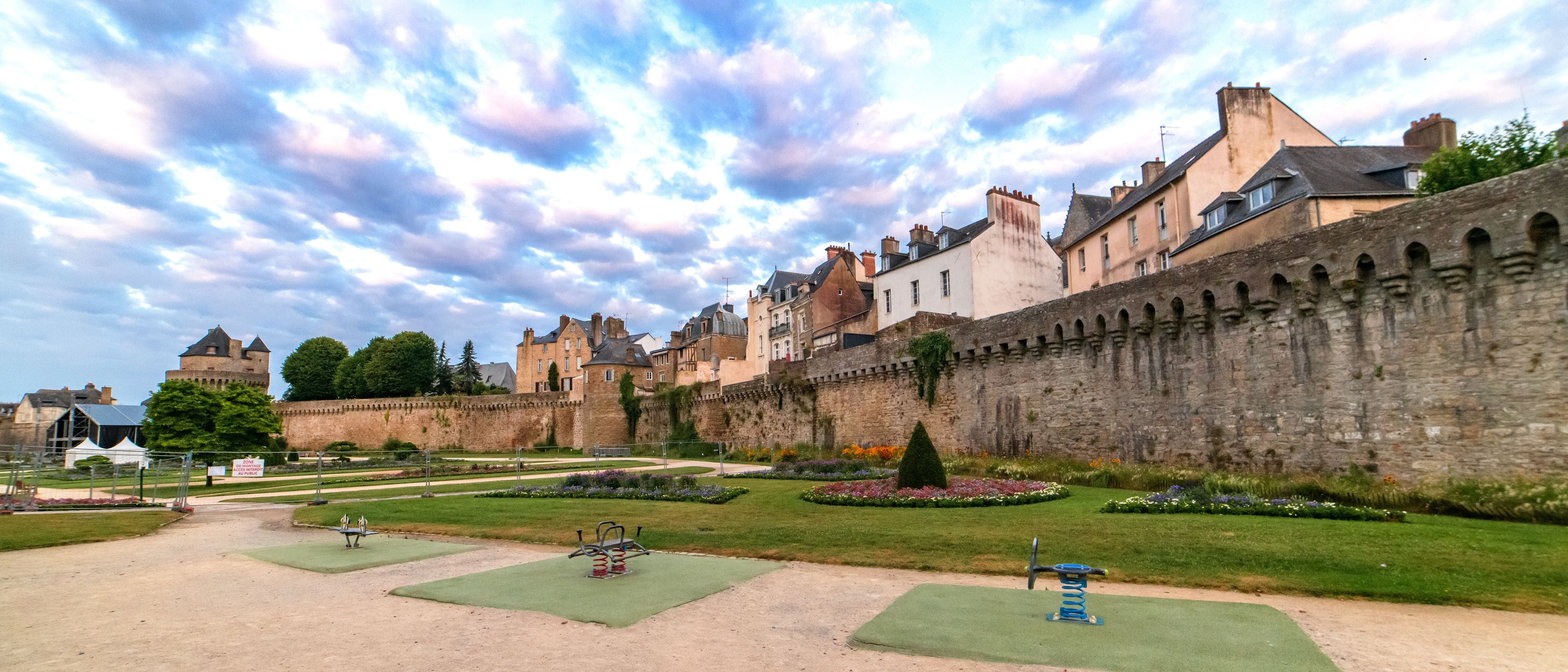 A historic castle with a large stone wall, a well-maintained garden with various plants and flowers, and a clear sky with clouds.